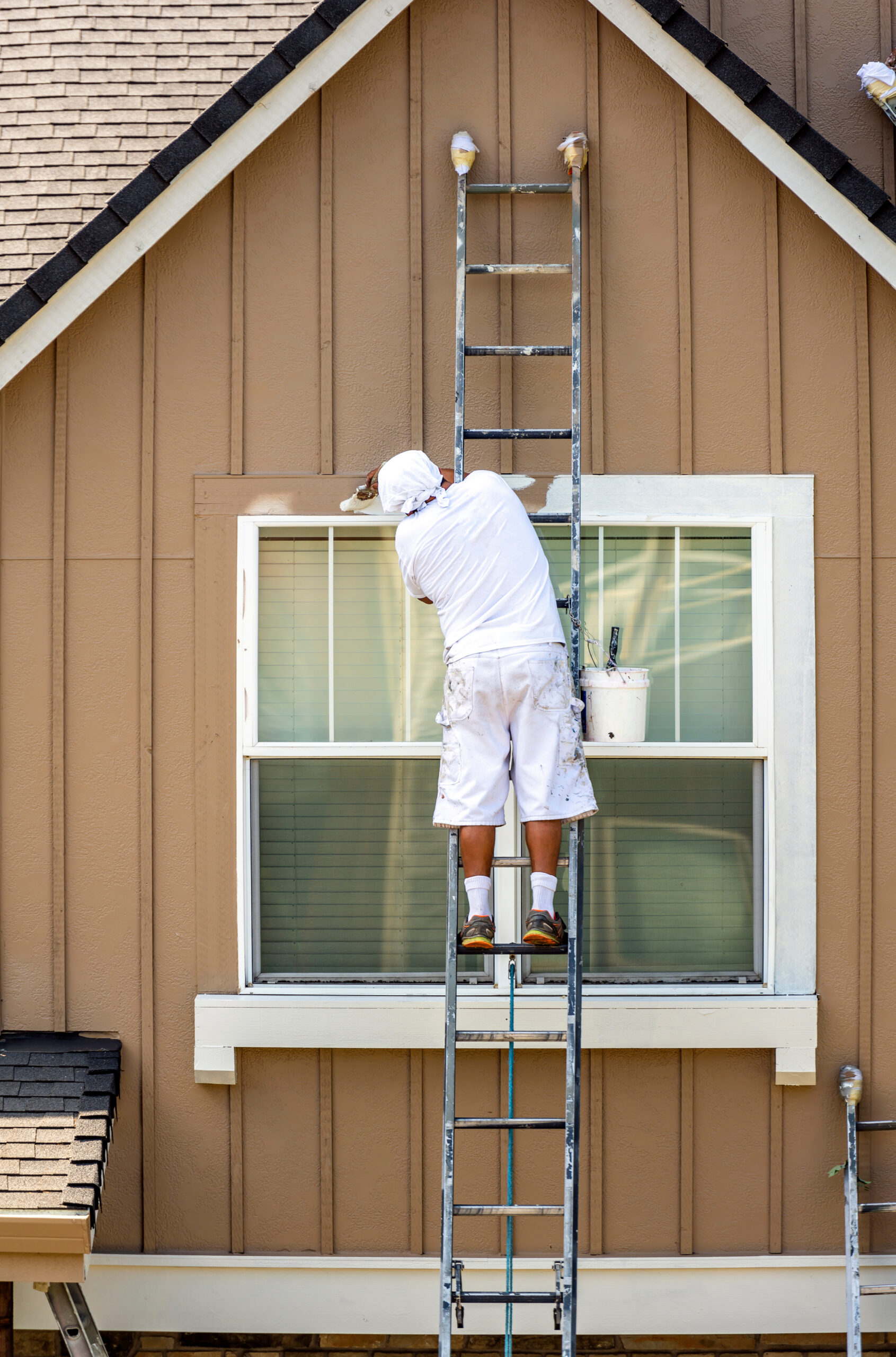 Painter man in white clothes with a protective scarf on his head paints a wooden window trim with a brush while standing on a high ladder leaning against the facade of a residential building